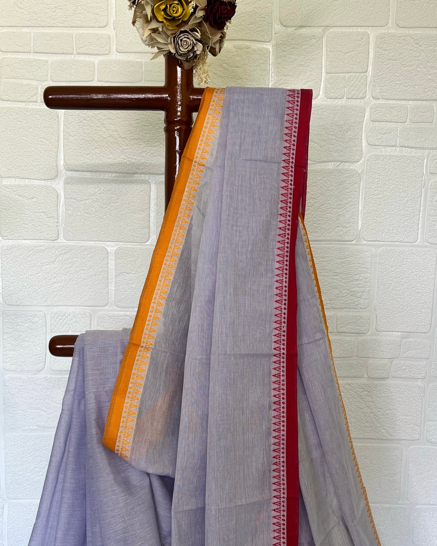 Gray khadi cotton saree with colorful borders draped over a wooden hanger against a white brick wall.