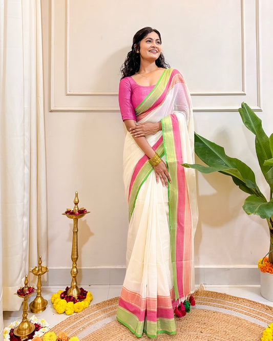 Woman wearing a white mulmul cotton saree with pink and green border in a decorated room.