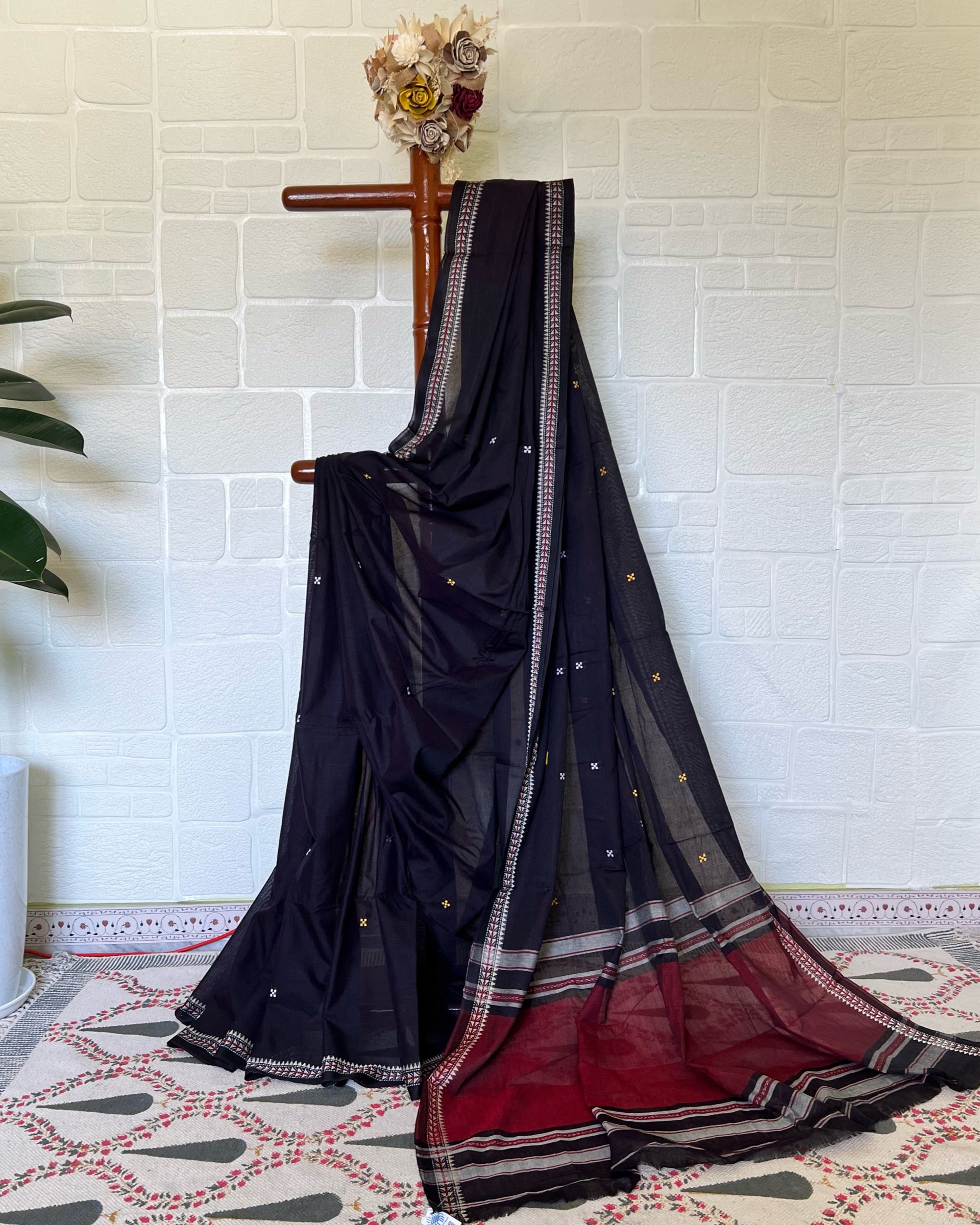 Black khadi cotton saree with red border draped over a wooden rod against a white brick wall.