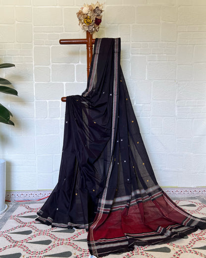 Black khadi cotton saree with red border draped over a wooden rod against a white brick wall.