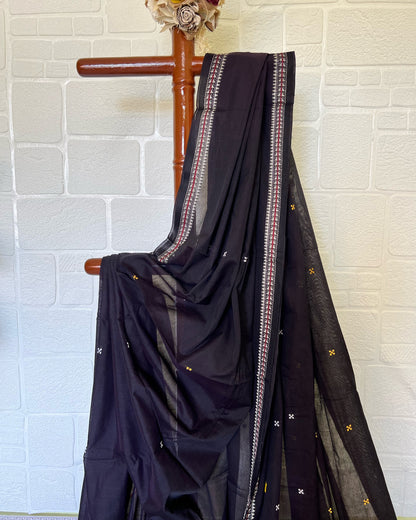 Black khadi cotton saree with white patterns draped over a wooden hanger against a light brick wall.