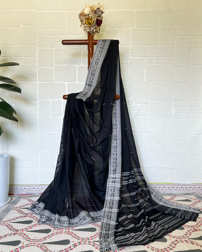 Black khadi cotton saree with white lace border draped over a wooden rod against a white brick wall.