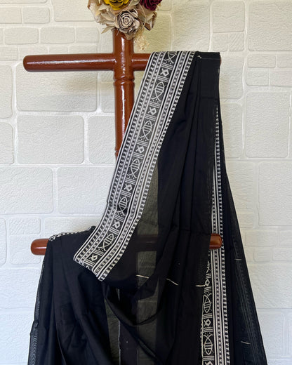 Black khaadi cotton saree with white embroidery draped over a wooden rod against a white brick wall.