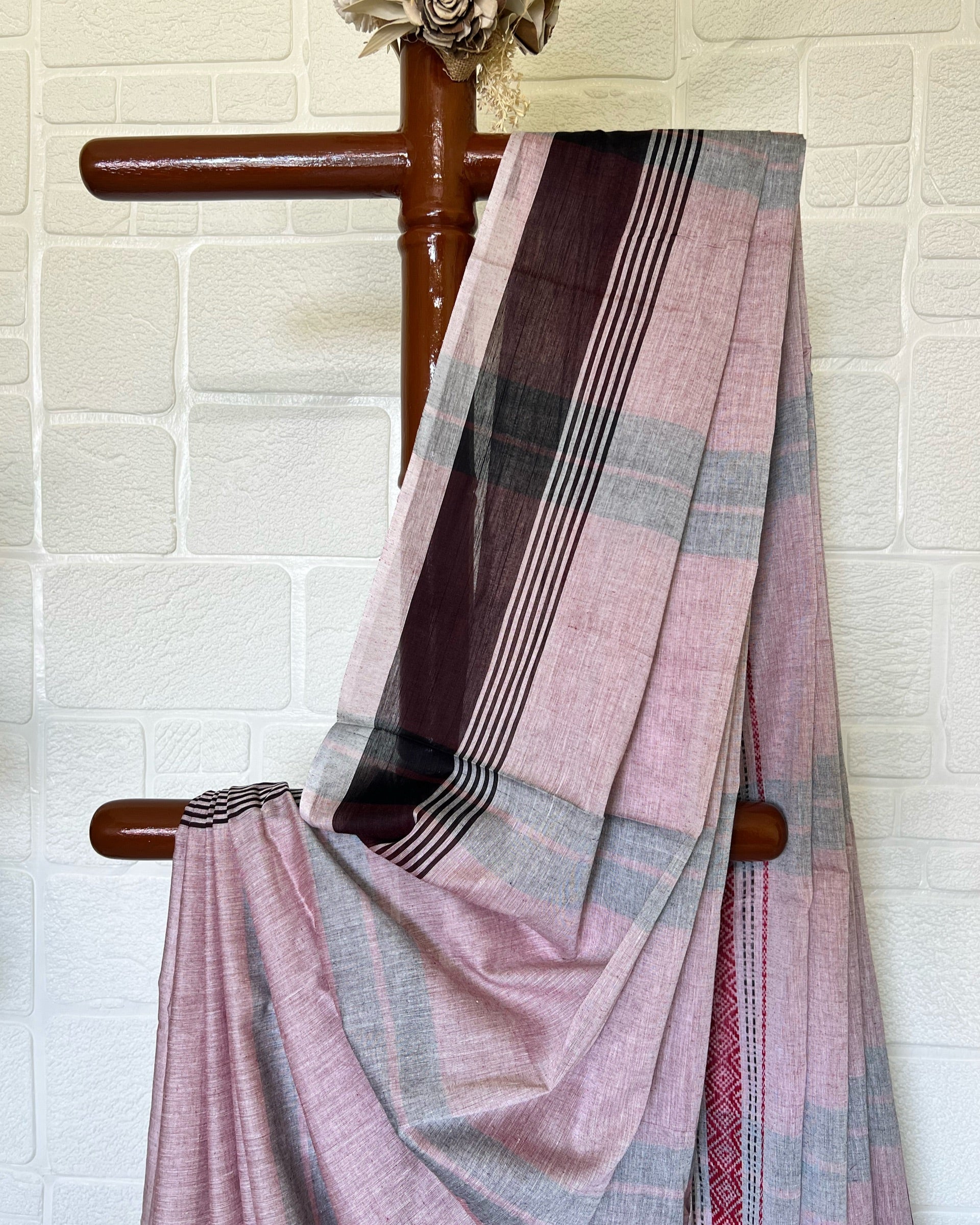 Pink and brown khadi cotton saree draped over a wooden chair against a white brick wall.