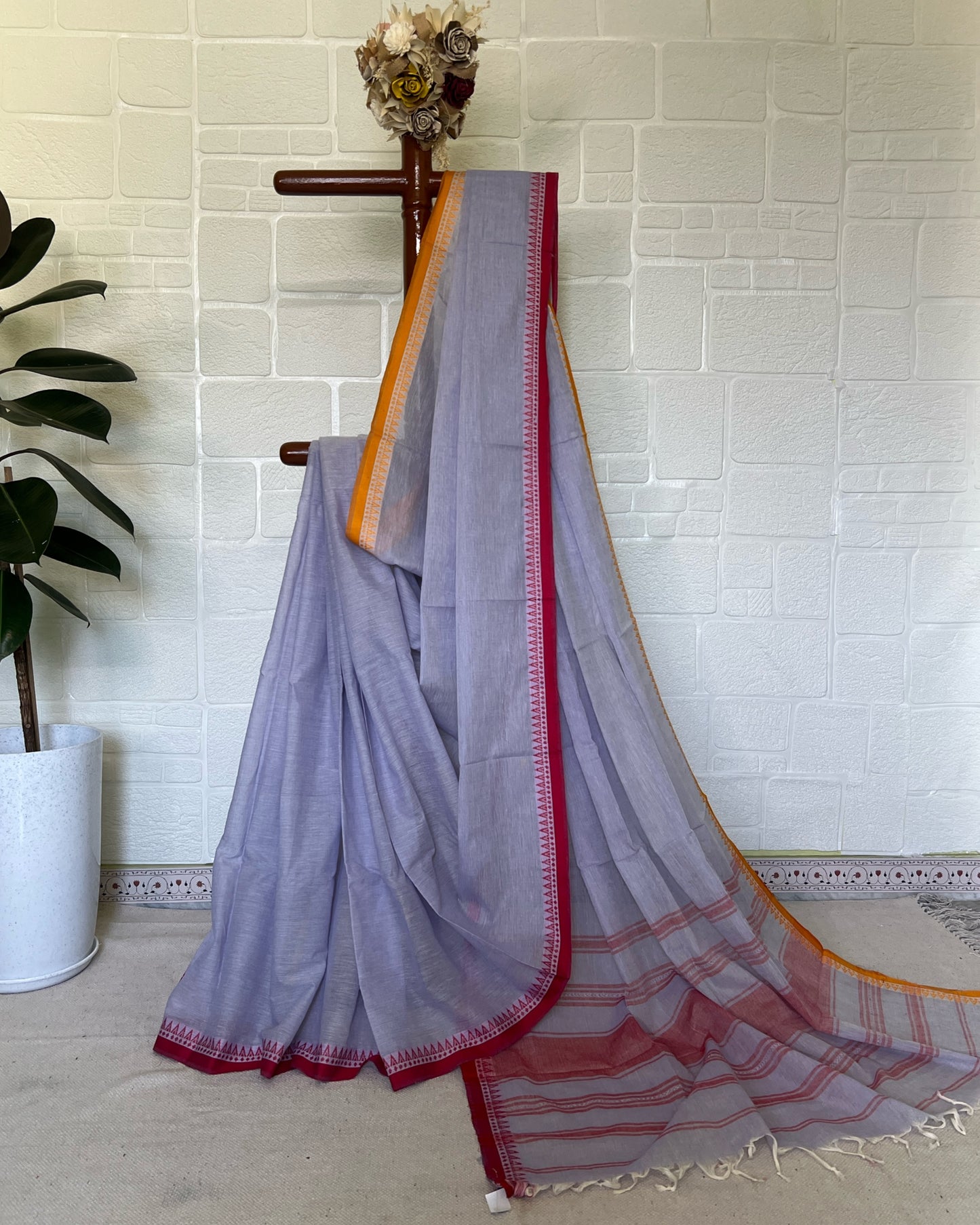 Light purple khadi cotton saree with red and orange border on a wooden hanger against a white tiled wall.