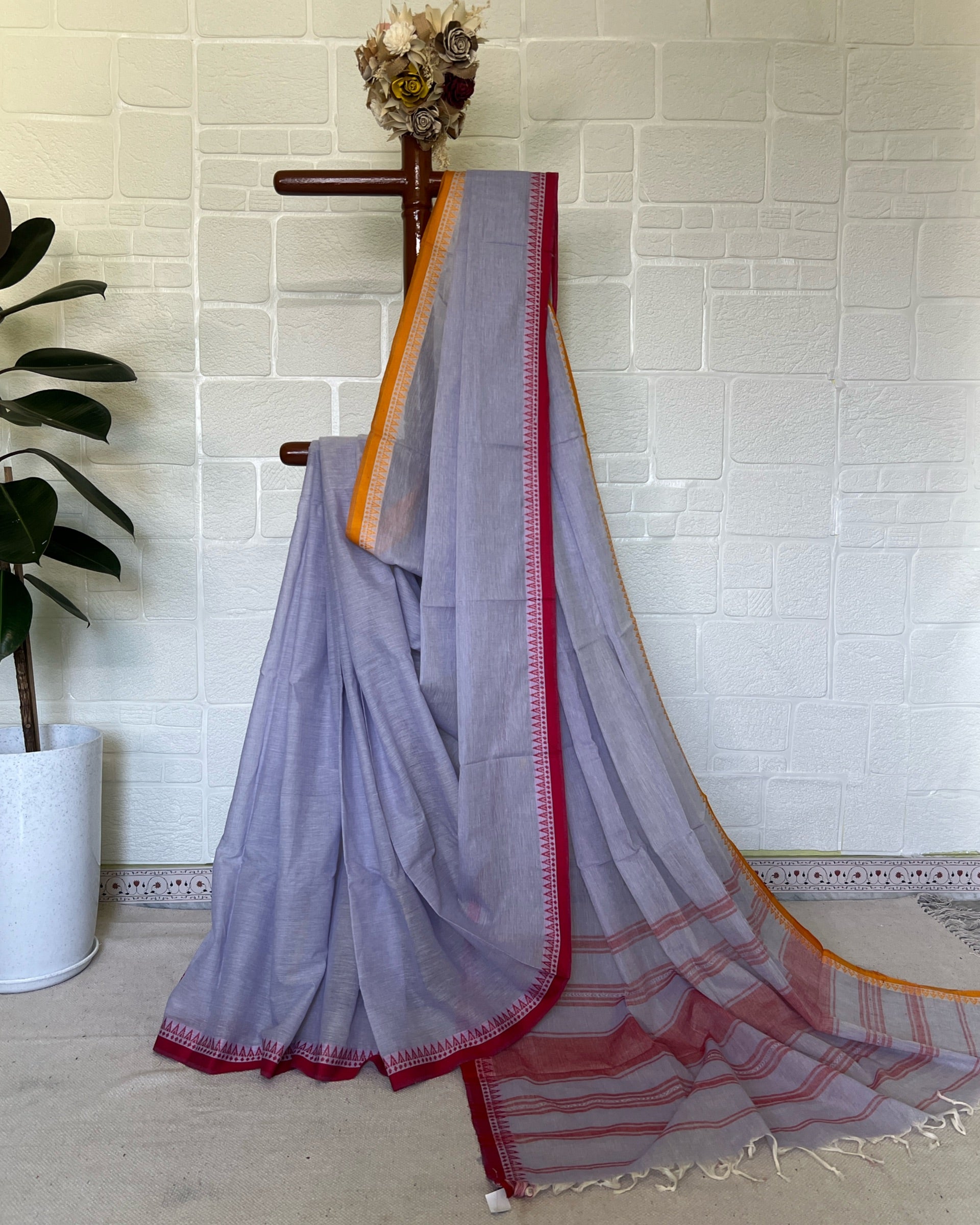 Light purple khadi cotton saree with red and orange border on a wooden hanger against a white tiled wall.