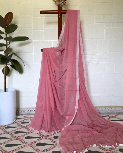 Pink handloom cotton saree draped over a wooden rod against a white brick wall.
