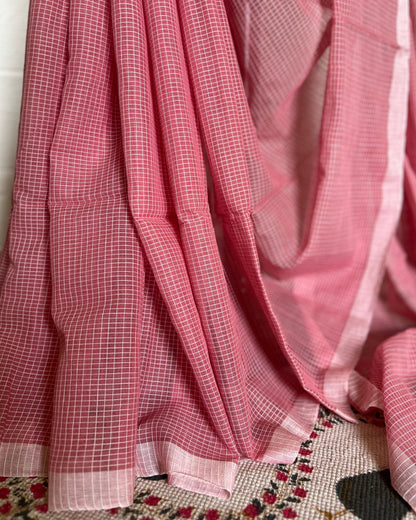 Pink checkered handloom cotton saree draped over a surface with a floral patterned rug underneath.