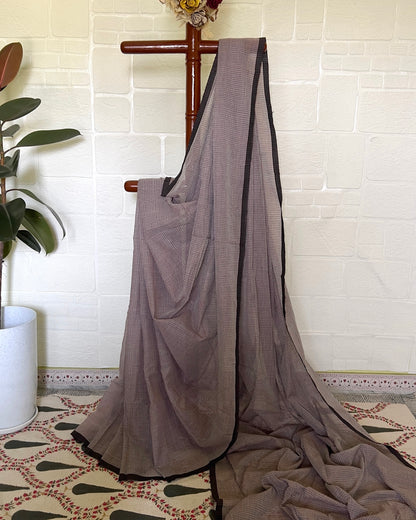 Brown handloom saree draped over a wooden hanger against a white tiled wall.