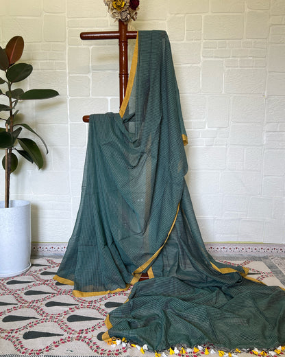 Green check handloom cotton saree draped over a wooden hanger against a white brick wall.
