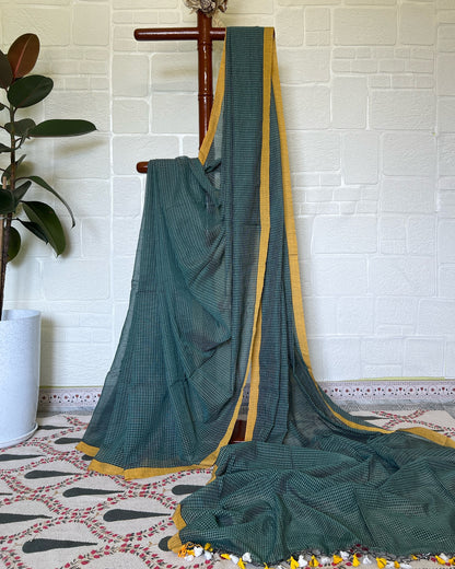 Green checkered handloom cotton saree draped over a wooden hanger against a white brick wall.