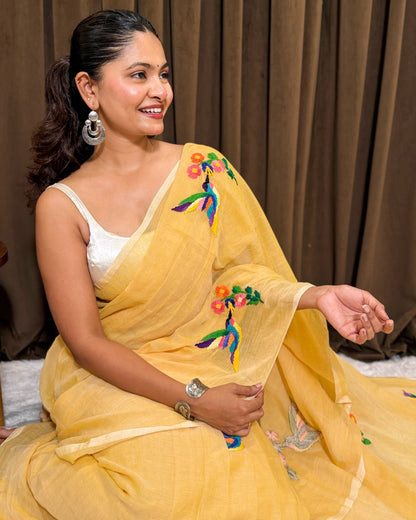 Woman wearing a yellow mulmul cotton saree with floral embroidery against a brown curtain background