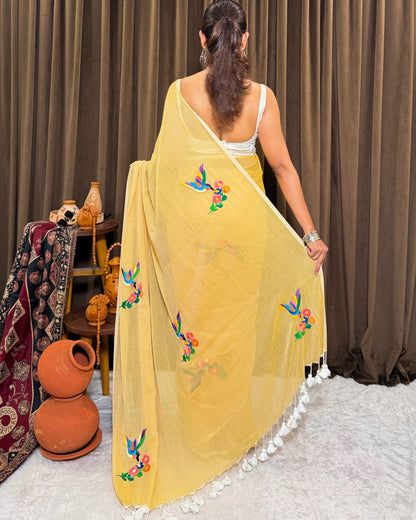 Woman wearing a yellow mulmul cotton saree with floral embroidery in a room setting.