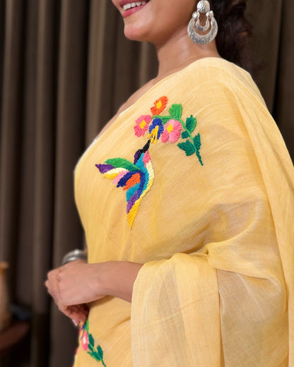 Woman wearing a yellow mulmul cotton  saree with floral embroidery, smiling against a brown curtain background.