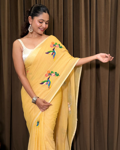 Woman wearing a yellow mulmul cotton saree by Shopaame with floral and bird embroidery against a brown curtain background