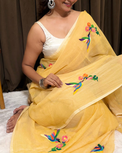 Woman wearing a yellow Shopaame mulmul cotton saree with floral embroidery against a brown curtain background