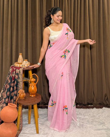 Woman in a pink mulmul cotton saree  by Shopaame with floral embroidery standing next to a small wooden table with decorative items against a brown curtain background.
