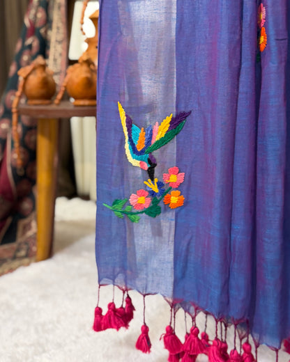 Purple embroidered mumul cotton saree by Shopaame with colorful bird design and pink tassels, set against a blurred indoor background.