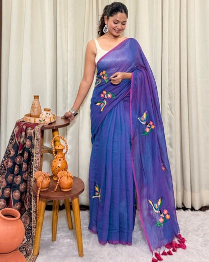 Woman wearing a purple mulmul cotton saree with floral embroidery in a room with decorative items.