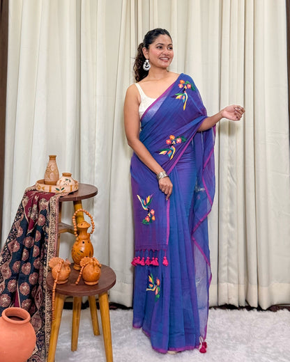 Woman wearing a purple mulmul cotton saree by Shopaame with floral patterns, standing next to a small wooden table with decorative items.