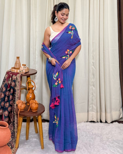 Woman wearing a purple mulmul cotton saree with floral patterns in a room with white curtains and decorative items.